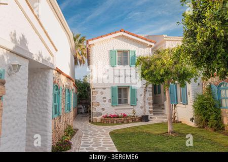 13. Mai 2026, Sigacik, Turkiye: Traditionelle ägäische Gästehausarchitektur mit türkisfarbenen Rollläden, gepflasterten Wegen und üppigem Garten Stockfoto