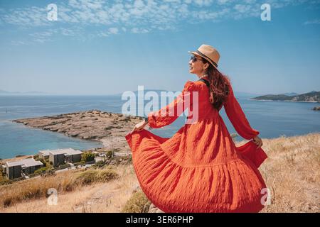 Frau in rotem Kleid, Sonnenhut und Sonnenbrille posiert auf einem Hügel und genießt während der Sommerreise den weiten Blick aufs Meer Stockfoto