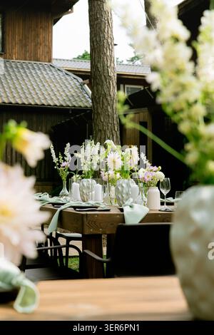 Langer Hochzeitstisch im Garten mit weißen und gelben Blumen, Blick durch Glastüren, dunkle Holzmöbel und Waldkulisse Stockfoto