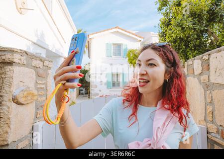 13. Mai 2026, Sigacik, Turkiye: Frau, die ein Smartphone hält, spricht und ein Live-Video aufnimmt, das ein charmantes weißes Haus während der Sommerreise zeigt Stockfoto