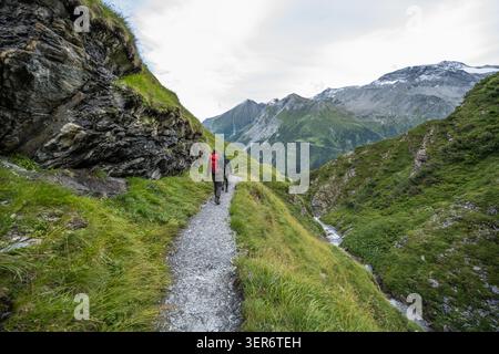 Zwei Erwachsene wandern einen Wanderweg in den Bergen an einem Fluss mit schneebedeckten Gipfeln Stockfoto