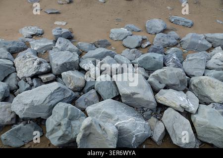Große graue Felsbrocken und verstreute Steine ruhen an einer Sandküste, die Natursteintexturen, weiße Adern und geologische Küstenformationen zeigen Stockfoto