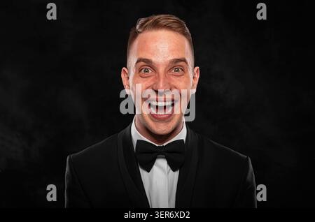 Ein fröhlicher junger Mann in einem formellen schwarzen Smoking und Bowtie lächelt aufgeregt mit weit geöffnetem Mund. Stockfoto