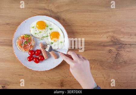 Spiegeleier mit Wurst, Kirschtomaten und Buchweizenbrot mit Lachsfisch. Mit den Händen im Rahmen Stockfoto