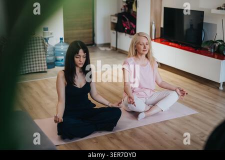 Zwei Frauen meditieren gemeinsam auf Yogamatten in einem hellen Wohnzimmer zu Hause Stockfoto