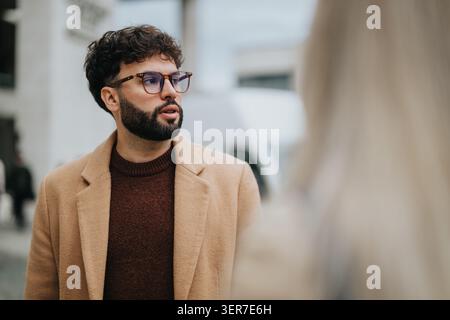 Junger Geschäftsmann in Kamelmantel und Brille, der auf einer städtischen Straße nachdenklich aussieht Stockfoto