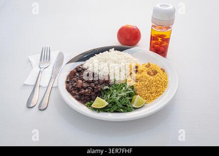 Feijoada, traditioneller brasilianischer Eintopf, serviert auf einem Teller mit Reis, Farofa, Gemüse, Limette, Tomaten, und Pfeffersoße. Ausgewogene Beleuchtung, leuchtende Farben, ein Stockfoto