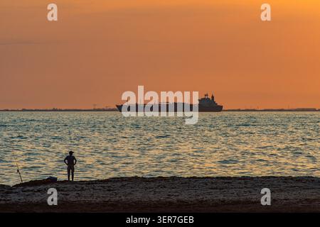 Fischer, die am Strand stehen und große Tankschiffe beobachten, die den Horizont bei Sonnenuntergang überqueren Stockfoto