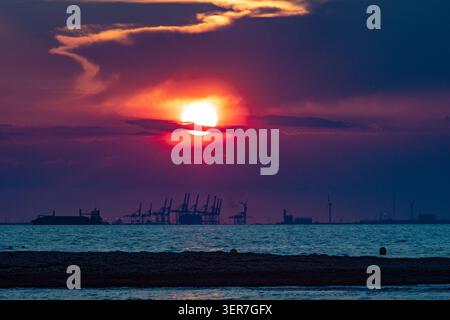 Die Sonne untergeht orange und rot über einem Industriehafen, mit Containerkränen und Frachtschiff Stockfoto