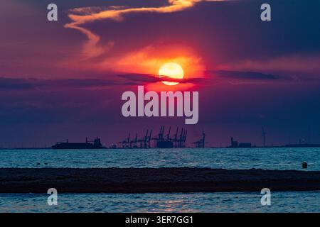 Sonnenuntergang beleuchtet einen Frachthafen mit Kränen und einem Schiff, reflektiert auf ruhigem Meerwasser Stockfoto