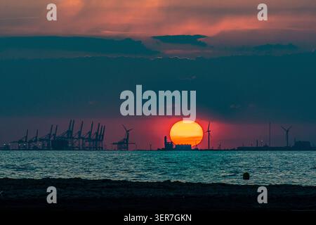 Sonnenuntergang beleuchtet Windturbinen und Containerkräne an einem Industriehafen Stockfoto