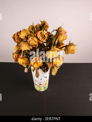 Goldene getrocknete Rosen füllen eine Blumenvase. Sie sind braun mit gelben Anklängen. Die weiße Vase hat bunte Muster. Hintergrund zeigt eine einfache Wand und dar Stockfoto