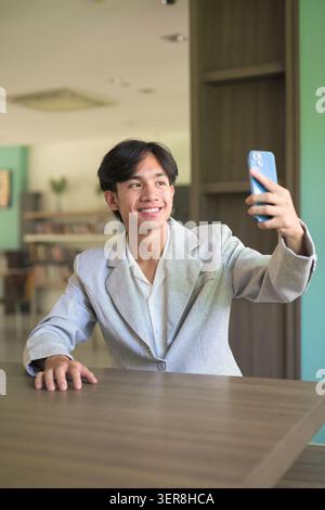 Authentisches vertikales Lifestyle-Porträt eines jungen, nicht-binären Thai-Isan-Profis oder Studenten, der beim Selfie lächelt. Halten Sie ein Smartphone für Stockfoto