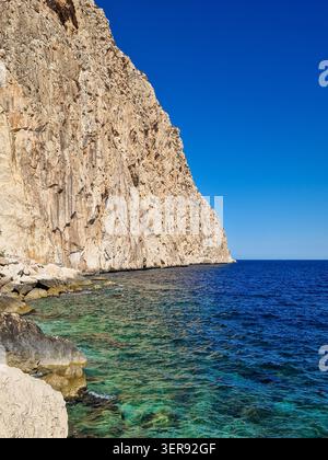 Sonnendurchflutete felsige Klippen fallen in kristallklares türkisfarbenes Mittelmeer, mit ruhigen Wellen, Unterwassersteinen und tiefblauem Horizont unter wolkenlosem Himmel Stockfoto