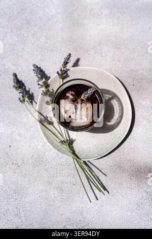 Ein erfrischendes Glas Eiskaffee mit Lavendelzweigen sitzt elegant auf einer Keramikplatte und fängt das Wesen des Sommers mit einem beruhigenden Fl ein Stockfoto