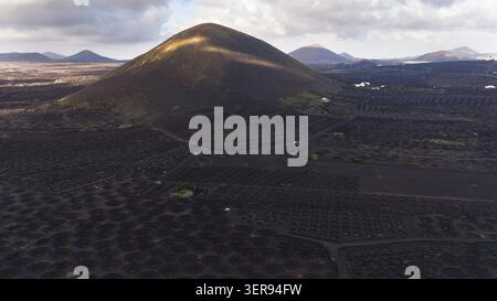 Magisches Licht schwingt über einen vulkanischen Kegel und schwarze Lavaberge auf Lanzarote, Kanarischen Inseln, mit Traubengruben und Steinmauern, die einen dramatischen, Stockfoto