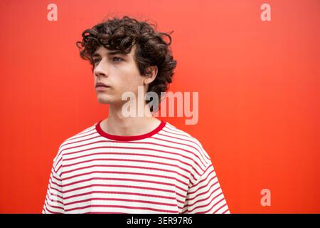 Ein junger Mann mit lockigen Haaren in einem rot-weiß gestreiften Hemd posiert im Profil vor einer leuchtend orangefarbenen Wand und vermittelt einen lässigen Lebensstil, Selbstvertrauen und Mod Stockfoto