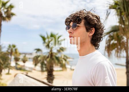 Seitenprofil eines lockigen Mannes mit Sonnenbrille und weißem T-Shirt, der draußen an einem sonnigen Strand mit Palmen steht und entspannte Summen einfängt Stockfoto
