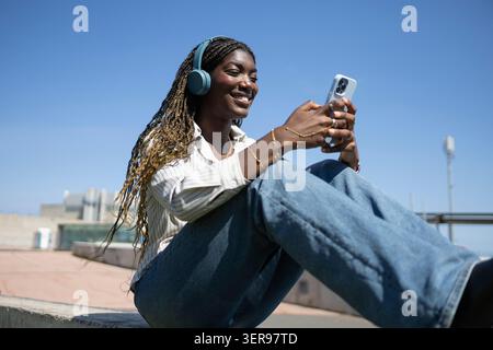 Eine Frau mit geflochtenen Haaren genießt es, Musik über kabellose Kopfhörer zu hören, während sie im Freien sitzt. Sie hält ein Smartphone und lächelt unter einer Clea Stockfoto