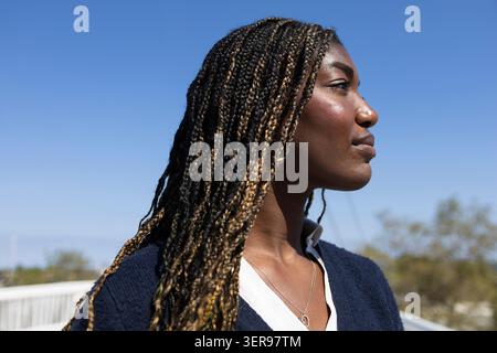 Eine Frau mit komplizierten Zöpfen steht selbstbewusst draußen und sonnt sich im Sonnenlicht. Das Bild unterstreicht ihren lebendigen, stylischen Look und zeigt ihren EM Stockfoto