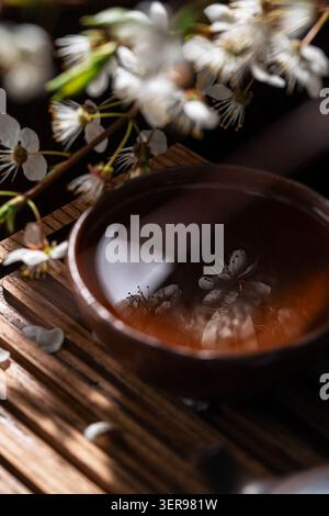 Dunkles, stimmungsvolles Stillleben der chinesischen Teezeremonie mit einer Ton-Teetasse auf rustikalem Holz, warmer Bernsteintee reflektiert zarte weiße Blüten, Ruhe und ich Stockfoto