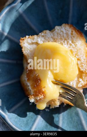 Nahaufnahme eines Lebensmittelfotos von süßem Zitronenquark, der über ein Butterkuchenstück auf einem blauen Teller tropft, mit einer Gabel. Helles Zitrus-Dessert-Konzept, hausgemachter Backofen Stockfoto