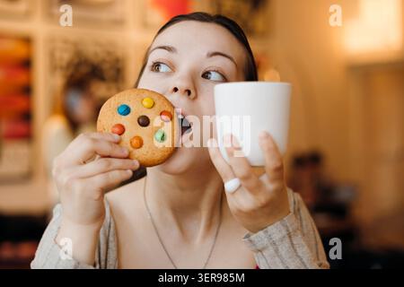 Nahaufnahme einer jungen Frau in einem gemütlichen Café mit einer weißen Tasse Kaffee und einem bunten Süßigkeiten-Keks und einer süßen Snackpause mit warmen, verschwommenen Ba Stockfoto