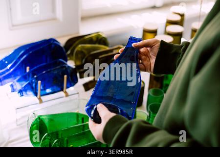 Eine Person hält ein leuchtend blaues Stück geschmolzenes Glas, das aus recycelten Flaschen hergestellt wird. Neben anderen farbenfrohen Stücken zeigt dieses umweltfreundliche Kunstemphas Stockfoto
