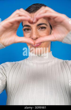 Eine Frau lächelt, die mit ihren Händen vor blauem Hintergrund eine Herzform macht, symbolisiert Fürsorge Stockfoto