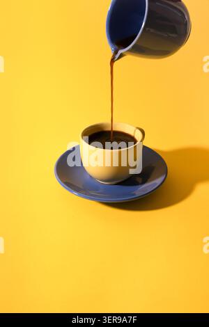 Schwarzer Kaffee wird aus einer kleinen Kanne in eine Keramikkkaffeetasse auf einer blauen Untertasse gegossen, die in einer minimalistischen Studioszene mit hellgelbem Hintergrund festgehalten wird Stockfoto
