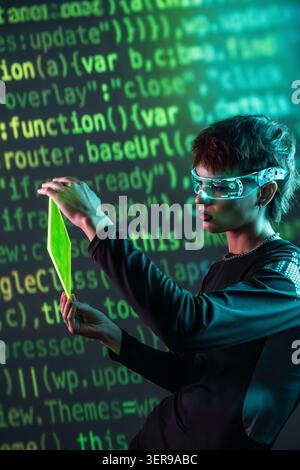 Futuristische junge Frau im Cyberpunk, die eine Virtual-Reality-Brille trägt, ein leuchtendes Panel vor grünem Code hält und ein Neon-digitales i erforscht Stockfoto