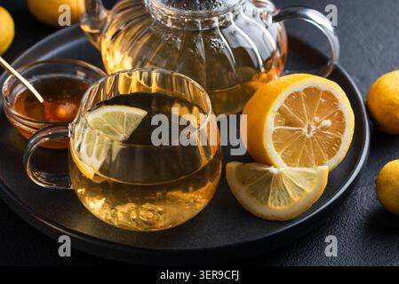 Teekanne aus klarem Glas und Tasse gefüllt mit goldenem Zitronentee, begleitet von frischen Zitronen und einer Schüssel Honig, präsentiert ein wohltuendes und gesundes Getränk Stockfoto