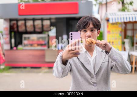 Authentisches horizontales Lifestyle-Porträt einer jungen, nicht-binären Thai-Isan-Person, die ein Selfie macht, während sie einen Trommelstock mit gebratenem Huhn hält. Content cre Stockfoto