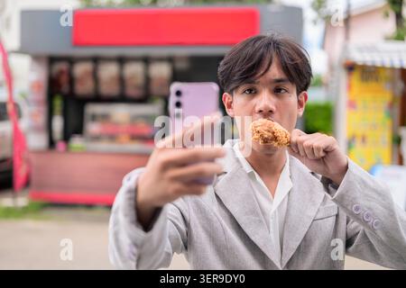 Authentisches horizontales Lifestyle-Porträt einer jungen, nicht-binären Thai-Isan-Person, die ein Telefon-Selfie macht, während sie einen Trommelstock mit gebratenem Huhn hält. Forts Stockfoto