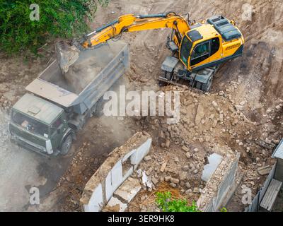 Die Ansicht von oben erfasst den Bagger, der Schutt auf der Baustelle in den Muldenkipper lädt. Staubwolken bilden sich, wenn schwere Maschinen unter Trümmern und Stockfoto