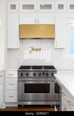 Eine weiße Kücheneinrichtung mit einem Marmorboden und einer Arbeitsplatte, einem Edelstahlofen und einer goldfarbenen Haube, Beschlägen und einem Wasserhahn. Stockfoto