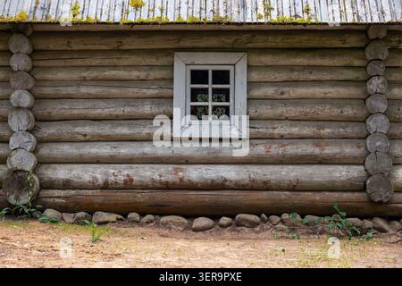 Verwitterte horizontale Stämme und ein kleines weiß gerahmtes Fenster an einer rustikalen Kabine außen, mit abblätternder Farbe, moosigem Metalldach und Steinfundament Stockfoto