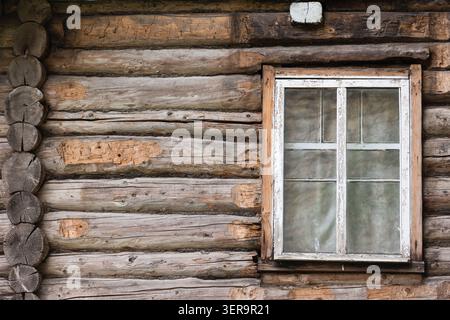 Eine verwitterte Holzblockhüttenwand mit einem abblätternden, weiß gerahmten Fenster mit Glasscheiben. Rustikale, Vintage-Textur und natürliche Holzmaserung sorgen für Hi Stockfoto