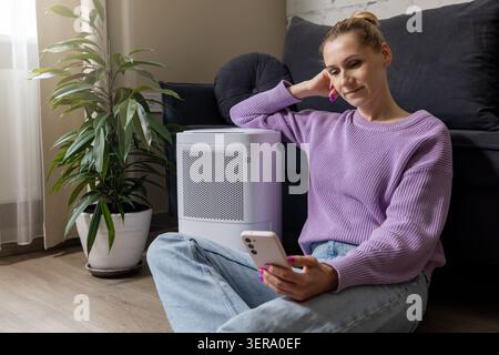Frau sitzt auf dem Boden mit Luftreiniger im Wohnzimmer und benutzt Handy. Luftqualität und Mikroklimaregelung zu Hause Stockfoto