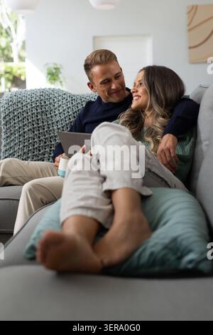 Diverse Paare sitzen dicht auf hellgrauem Sofa zu Hause, Mann mit Tablette, Frau mit Becher Stockfoto