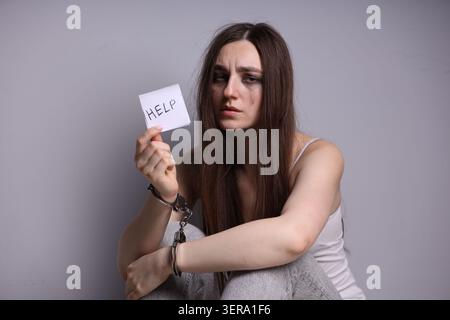 Menschenhandel. Frau mit Handschellen, die Papier mit Worthilfe auf grauem Hintergrund hält Stockfoto