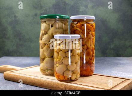 Drei Gläser mit verschiedenen Arten eingelegter Pilze. Darunter kleine Pilze und wilde Honig-Agaren. Auf einer rustikalen Holzschneide stehen Stockfoto