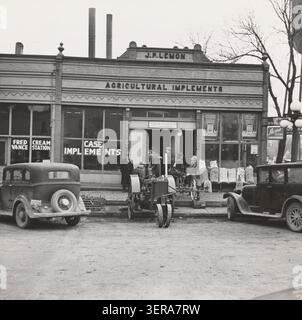 Historisches Foto einer Geschäftsstraße in Aledo, Illinois, mit einem Händlerbetrieb für landwirtschaftliche Geräte, einem Case-Traktor, frühen Automobilen, Werbespots und Einwohnern während der Zeit der Großen Depression. Fotografiert von Russell Lee im März 1937. Stockfoto