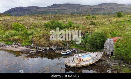 Eine ruhige Küstenlinie von Highland, an der verwitterte Boote, ein altes Bootshaus und zerklüftete Hügel die zeitlose Schönheit Schottlands einfangen Stockfoto