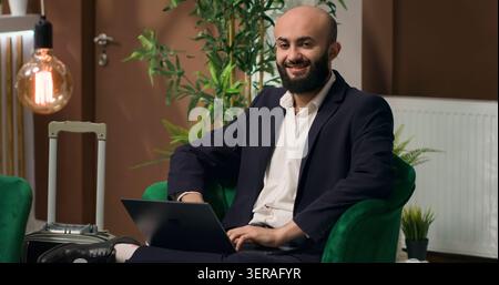 Ein Mann aus dem Nahen Osten tippt auf einem Laptop, während er sich im Lobbybereich entspannt und eine Rede für eine globale Firmenveranstaltung schreibt. Geschäftsreisender, Hotelrezeption. Stockfoto