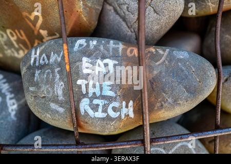 Ein handgemalter Flussstein mit der Aufschrift „Eat the Rich“ in einer Gabionenwand, Christchurch, Neuseeland, November 2025. Stockfoto