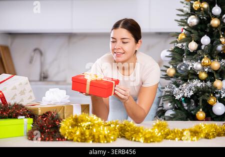 Fröhliche junge Frau in lässiger Kleidung, die ein Geschenkpapier neben dem Weihnachtsbaum im Wohnzimmer hält Stockfoto
