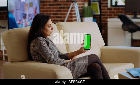 Person, die während der Arbeit von zu Hause aus E-Mails auf dem Green-Screen-Handy liest, indische Frau in gemütlicher Wohnung, die Aufgaben erledigt, Geschäftsnachrichten auf dem Mockup-Handy liest, Schwenkschuss. Stockfoto