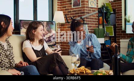 Kulturell inkludierte Gruppe von Frauen, die sich unterhalten, Wein trinken, über ihre Freunde tratschen, asiatische, afroamerikanische und kaukasische Mädchen, die das soziale Leben im Wohnzimmer genießen. Stockfoto