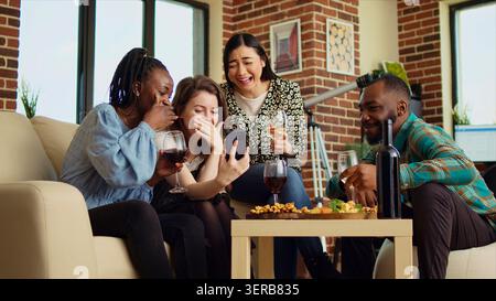Multiethnische Freunde beim Wiedersehen sehen sehen sich lustige Videos aus ihrer Kindheit auf dem Smartphone-Bildschirm an, fröhliche Gäste im Wohnzimmer sehen sich Clips von ihrer Jugend auf dem Handy an. Stockfoto